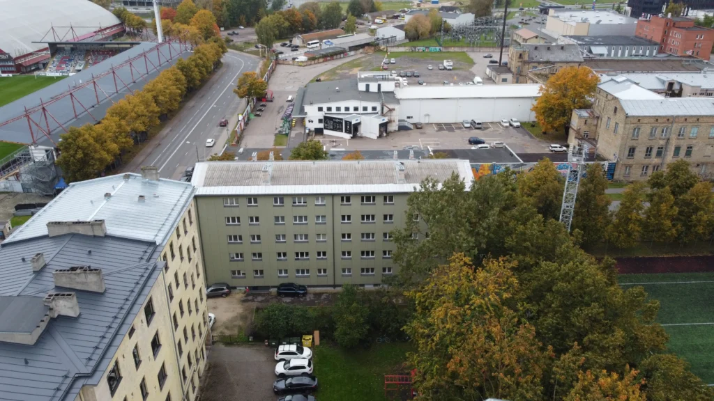 5 stāvu daudzdzīvokļu ēka ar zaļu fasādes apdari un gaišu jumtu, pie ēkas autostāvvieta un koki, Rīga