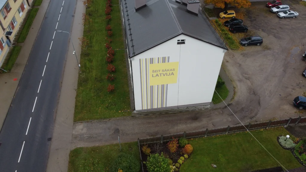 Renovēta daudzdzīvokļu māja ar baltu gala fasādi un tumši pelēku slīpu jumtu.