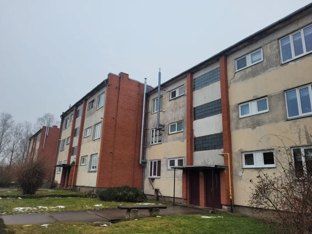 Daudzdzīvokļu ēka pirms atjaunošanas ar bēšu fasādi un sarkanu ķieģeļu kāpņutelpas blokiem.