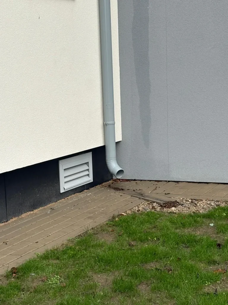 building facade detail with a rainwater downpipe and a ventilation grille near the base.