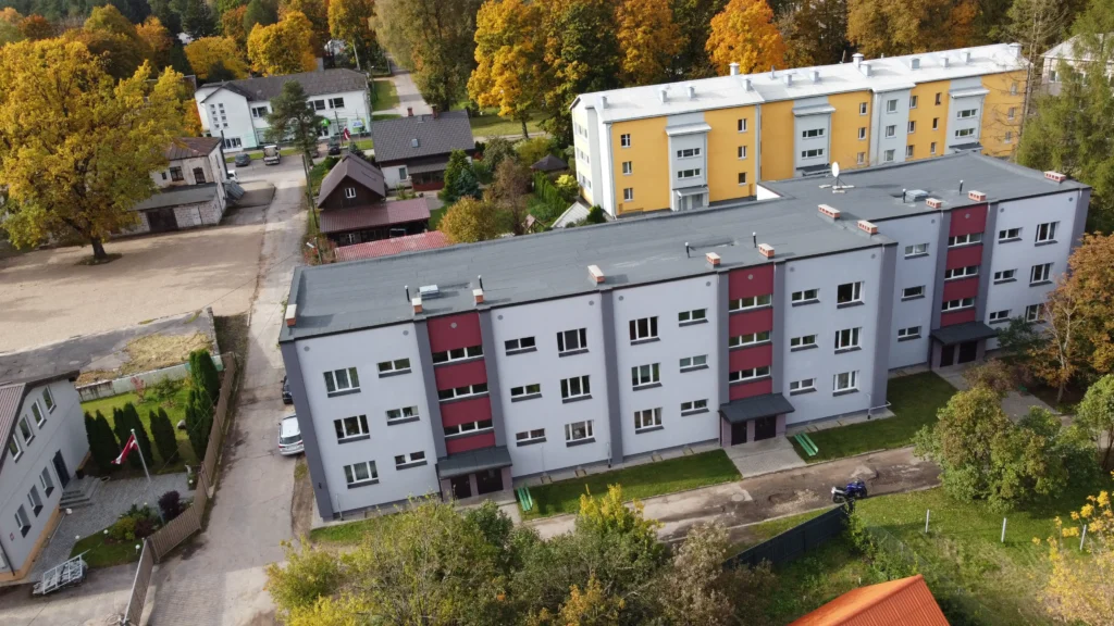 Daudzdzīvokļu ēkas piecstāvu nams ar sarkaniem fasādes akcentiem, fonā ar baltu un dzeltenu fasādes apdari un pelēku jumtu piecstāvu nams ar sarkaniem fasādes akcentiem