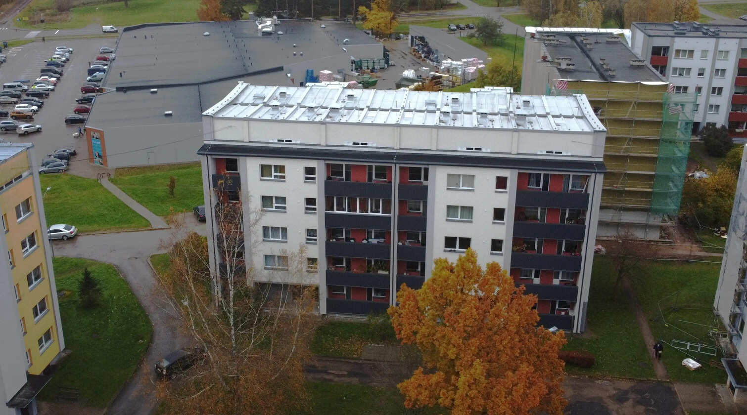 5 stāvu daudzdzīvokļu ēka ar gaišu fasādi, tumšiem balkoniem un pelēku jumtu.