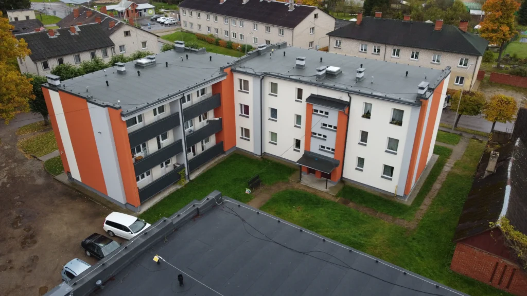 3 stāvu daudzdzīvokļu ēka ar baltu fasādi, oranžiem akcentiem, tumšiem balkoniem un plakanu tumšu jumtu