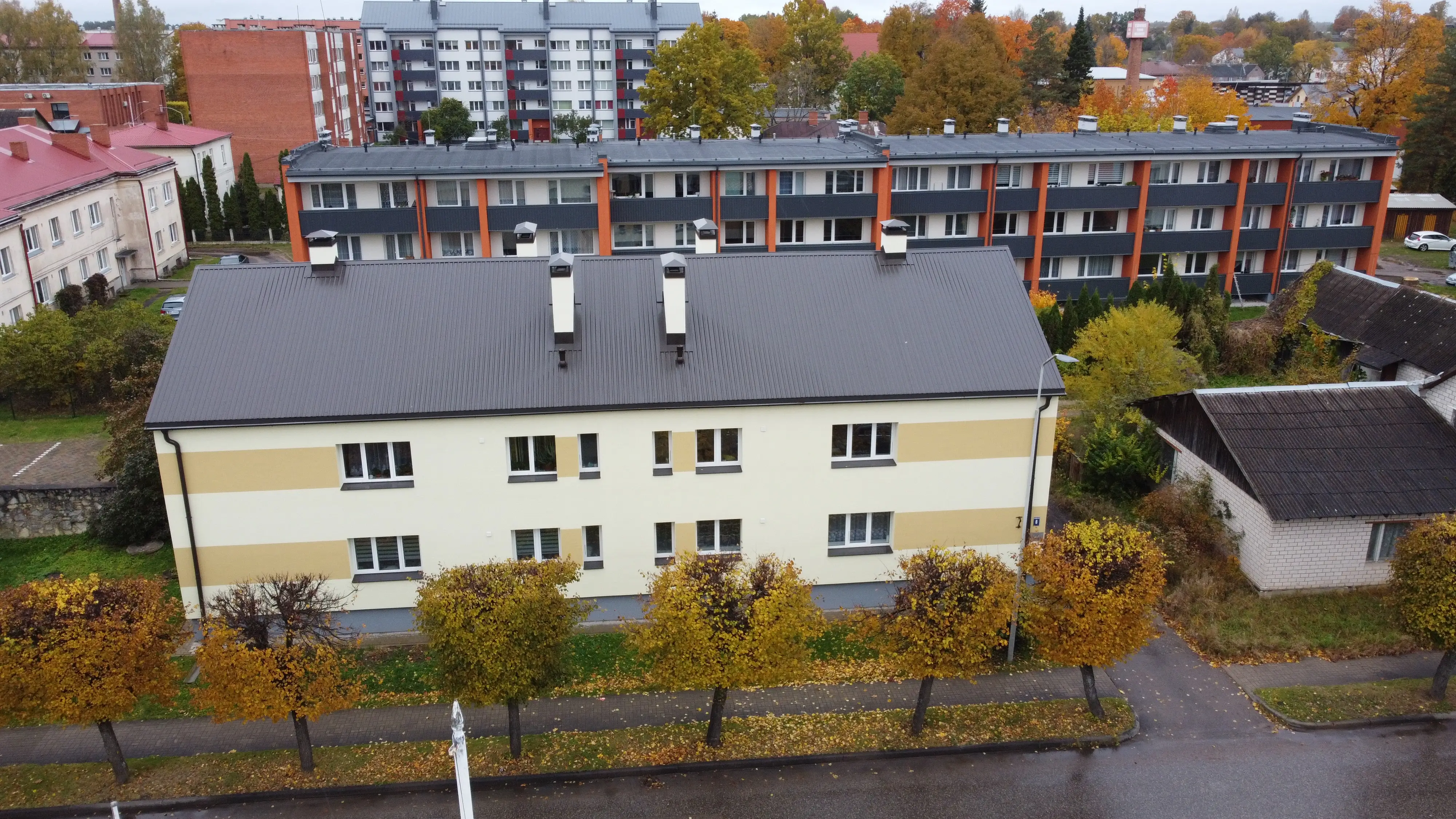 Divas daudzdzīvokļu ēkas: priekšplānā gaiša fasādes apdare ar tumšu jumtu, fonā ēka ar oranžām vertikālām joslām un balkoniem, Valka