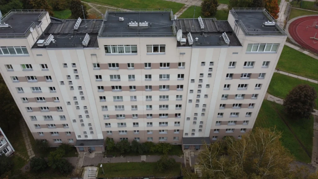 Renovēta 9 stāvu daudzdzīvokļu ēka ar gaiši bēšu fasādi, horizontālām joslām un tumšu plakanu jumtu.