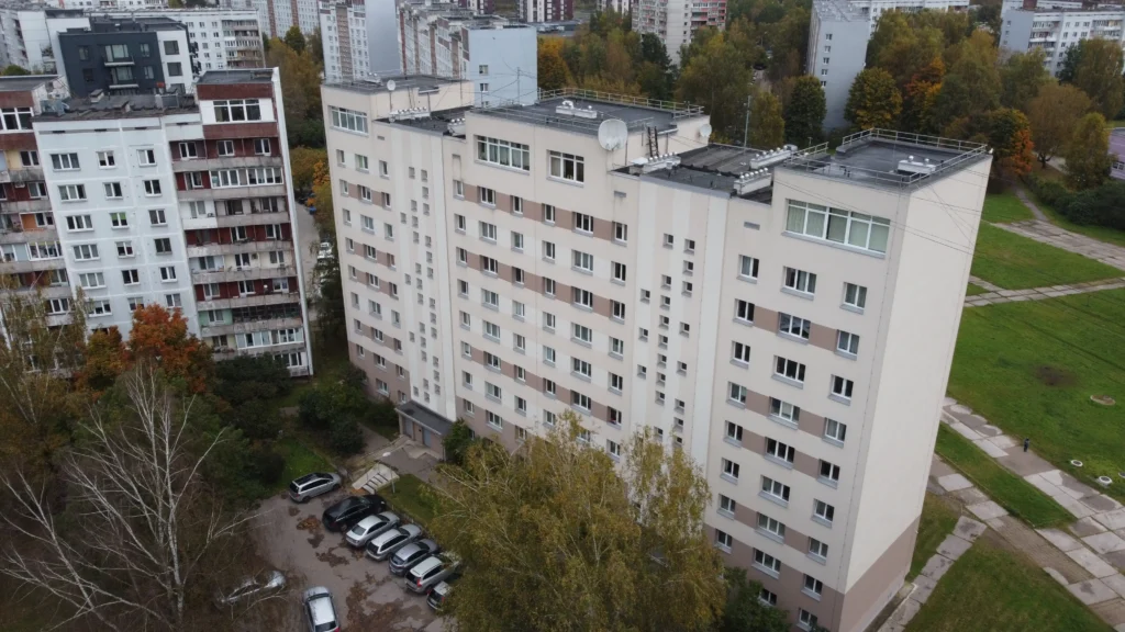 9 stāvu daudzdzīvokļu ēka ar baltu un pelēku fasādi, vienmērīgām logu rindām un melnu plakano jumtu