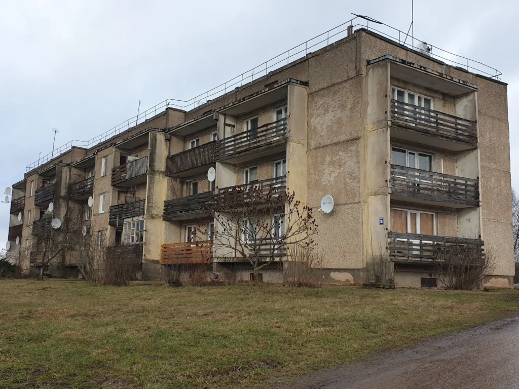 3 stāvu daudzdzīvokļu ēka ar nolietotu pelēko fasādi un veciem balkoniem