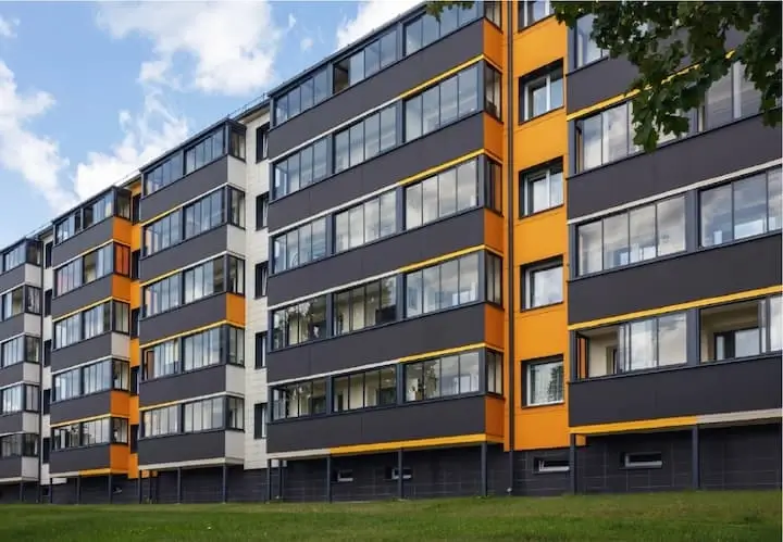 Renovēta daudzdzīvokļu māja ar pelēku fasādi, oranžiem akcentiem un stiklotajiem balkoniem