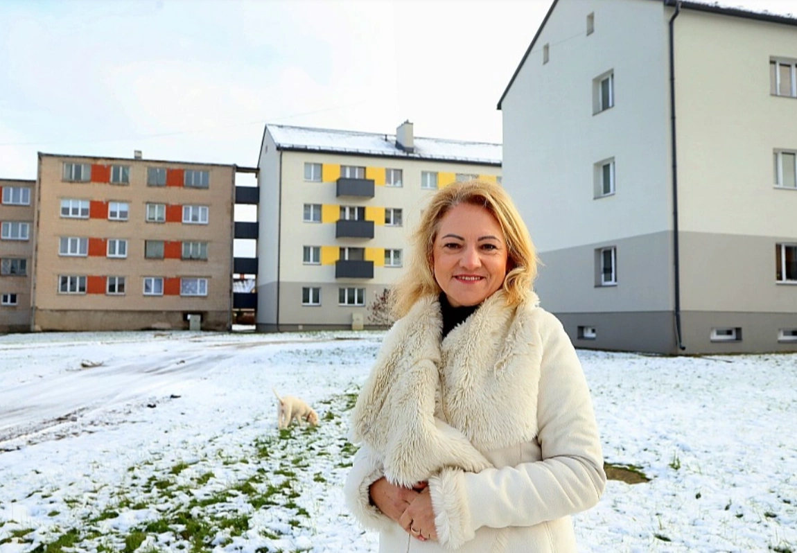 Sieviete pagalmā pie daudzdzīvokļu ēkām, fonā renovēta ēka ar dzelteniem akcentiem